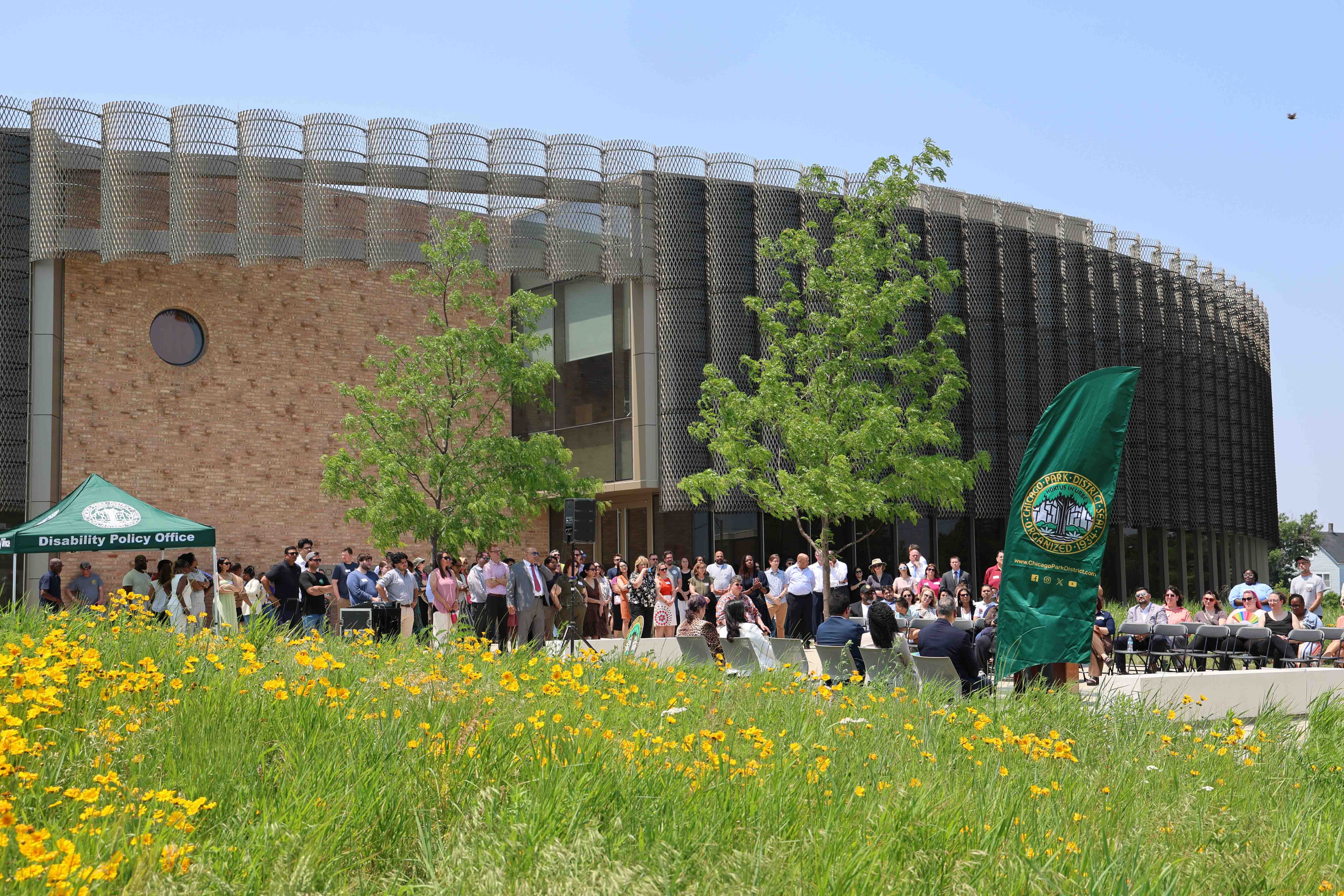 Crowd gathers outside a modern brick building with a unique metal facade, a "Disability Policy Office" tent, and a Chicago Park District banner. Yellow wildflowers are in the foreground.